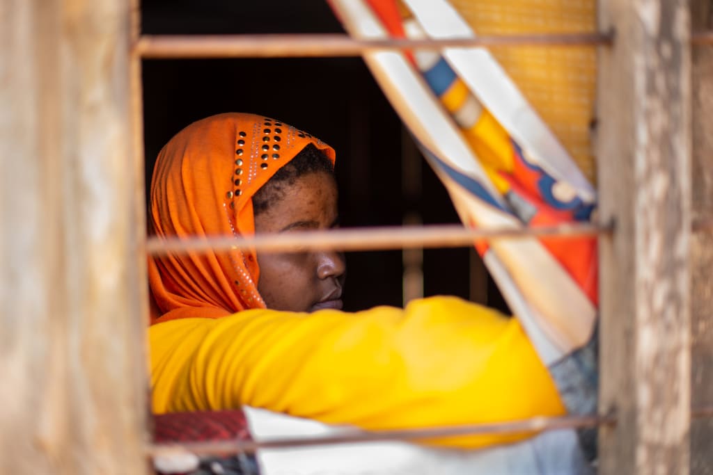 Sarah is wearing an orange head covering and is sitting in her grandmother's house. She is near the window.